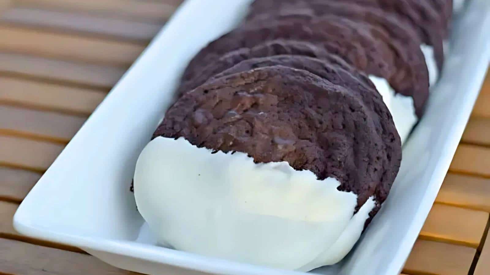 A row of chocolate cookies partially dipped in white frosting, arranged on a rectangular white plate on a wooden surface.