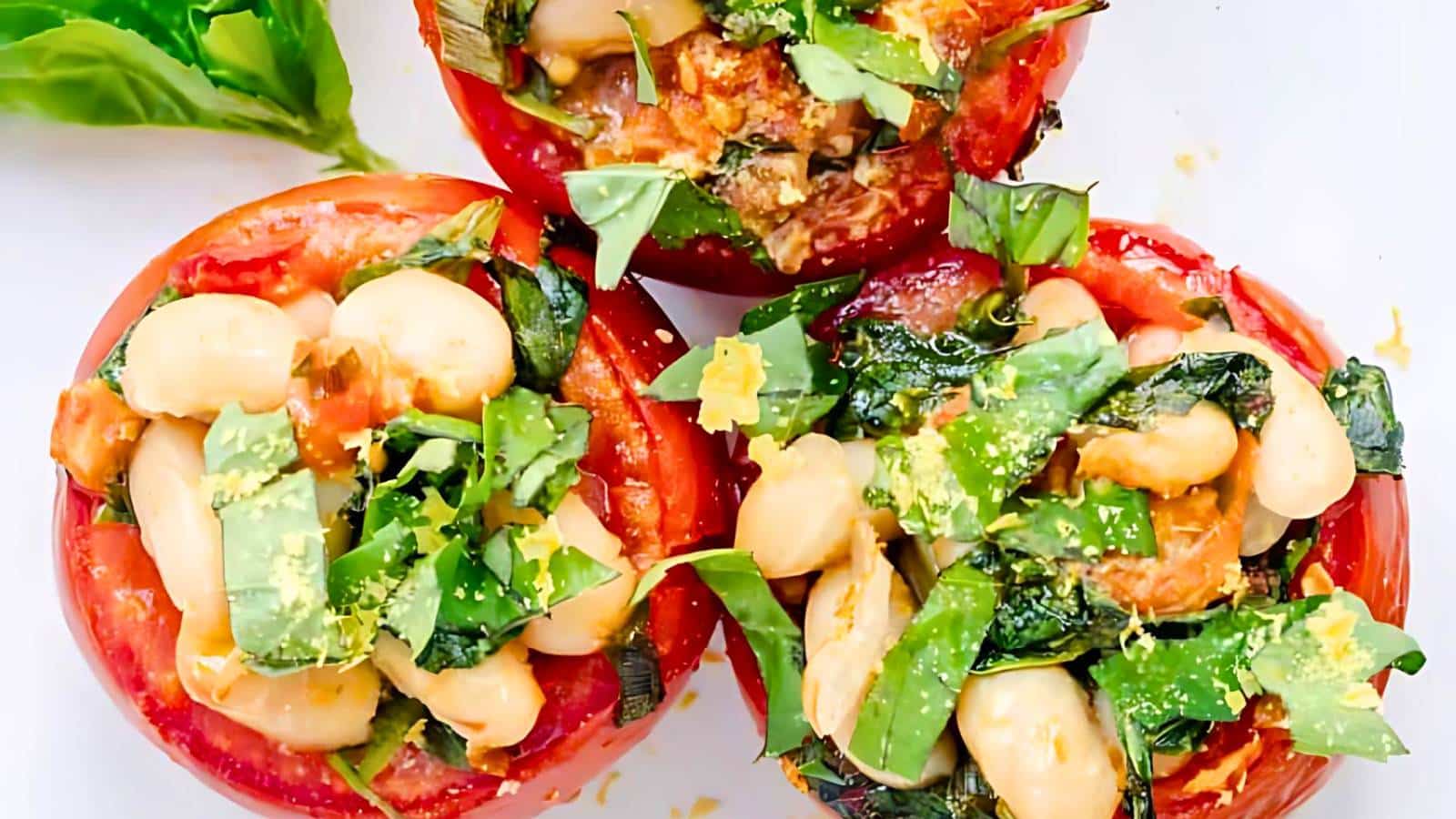 Three stuffed tomatoes filled with white beans, chopped greens, and garnished with lemon zest, displayed on a white surface.