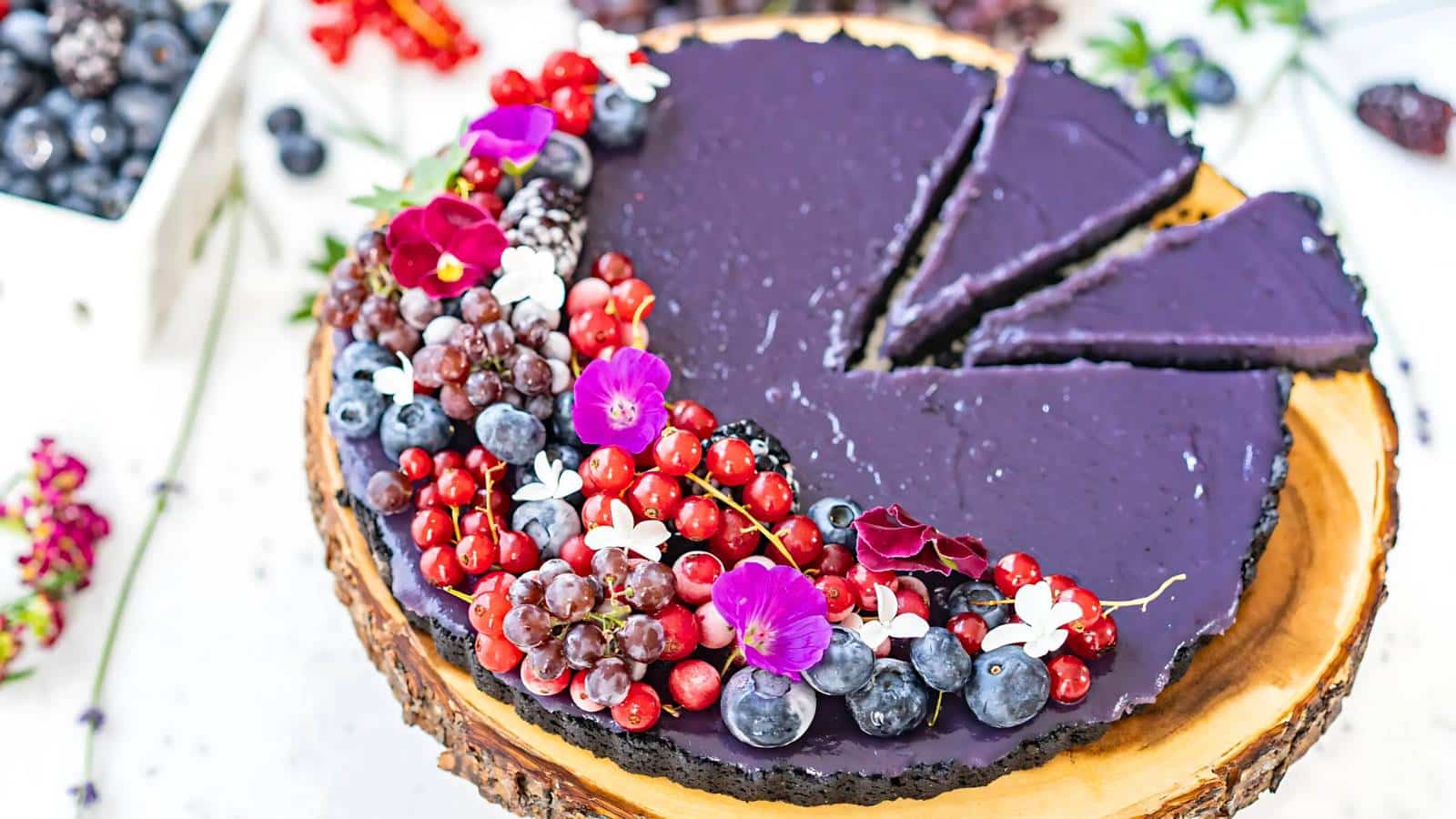 A purple tart on a wooden platform, garnished with a variety of fresh berries and edible flowers. Two slices are cut from the tart.