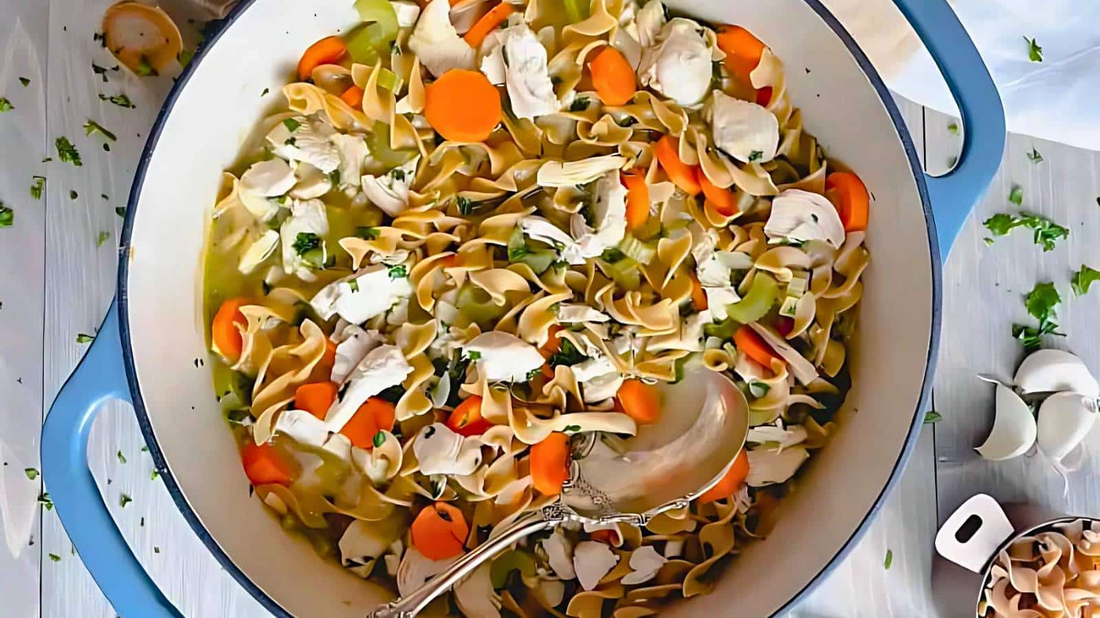 A blue pot filled with chicken noodle soup containing egg noodles, sliced carrots, celery, and parsley, with a silver ladle.