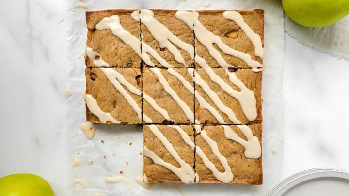 Nine apple blondies drizzled with icing are arranged on parchment paper, with two green apples nearby on a marble surface.