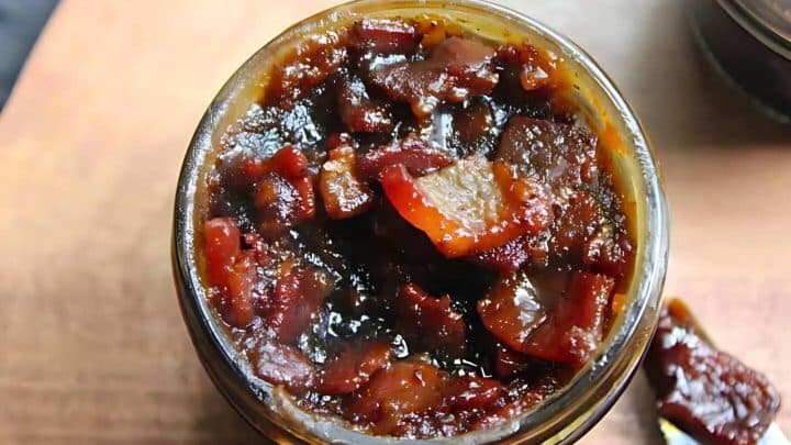Jar filled with bacon jam on a wooden surface, with visible chunks of bacon and a glossy texture.