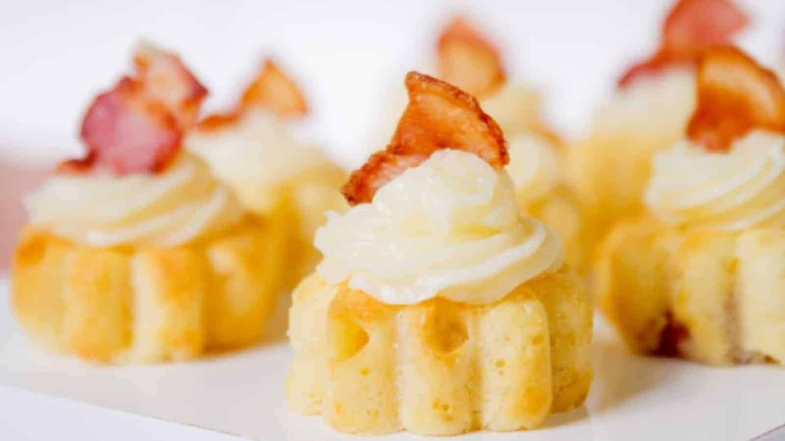 Mini cakes topped with creamy frosting and crispy bacon pieces on a white surface.