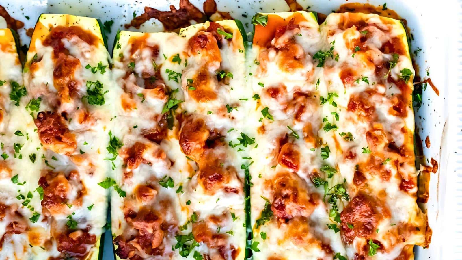 A close-up view of zucchini boats filled with a meat sauce, topped with melted cheese and garnished with chopped herbs.
