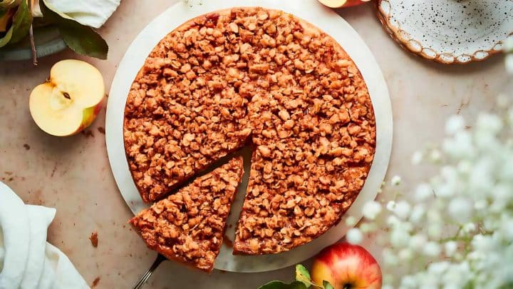 A round crumb-topped apple pie on a white plate with a single slice missing. Two halved apples and a plate are nearby on a marble surface.