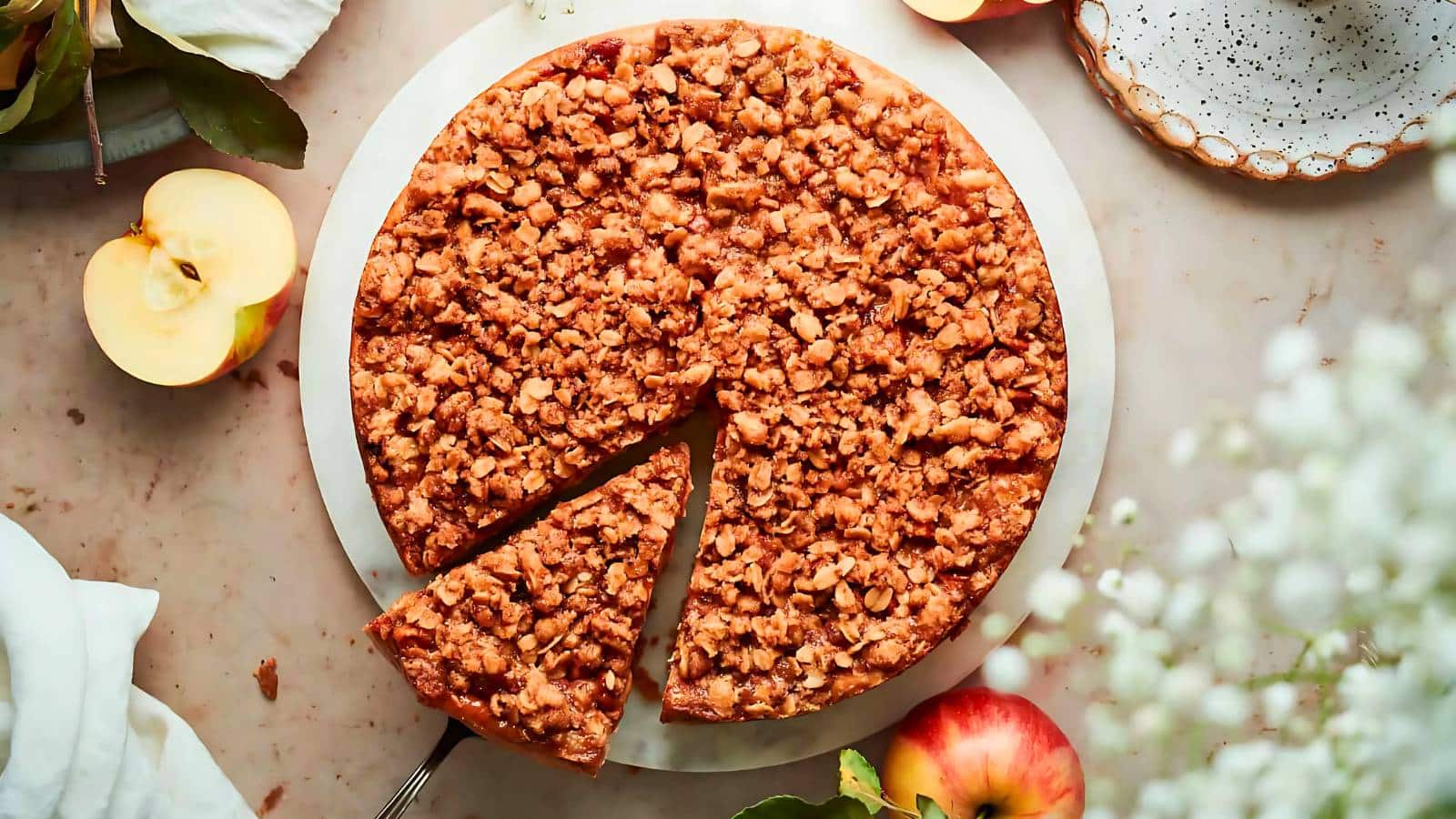A round crumb-topped apple pie on a white plate with a single slice missing. Two halved apples and a plate are nearby on a marble surface.