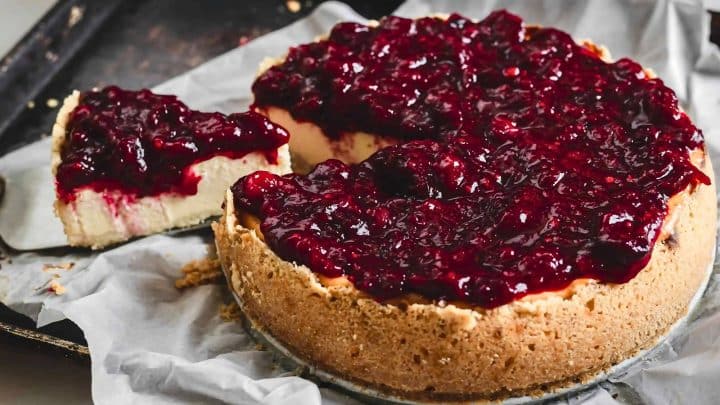 A cheesecake topped with a glossy layer of berry compote sits on parchment paper, with one slice cut out and placed nearby.