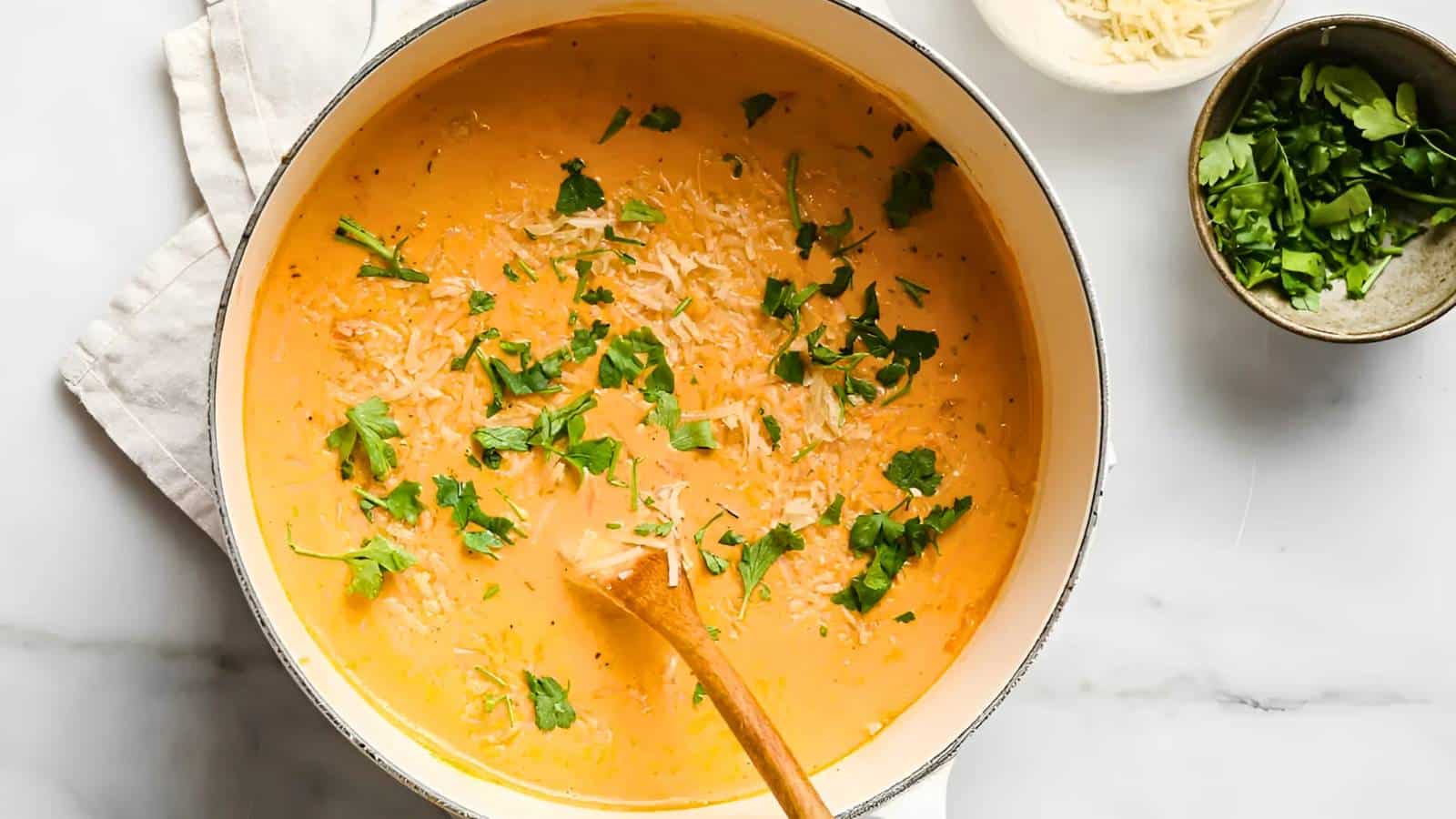 A pot of creamy orange soup garnished with chopped herbs, accompanied by small bowls of grated cheese and more herbs on the side.