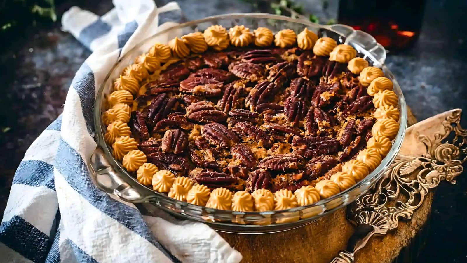 A pecan pie, one of the yummy Thanksgiving desserts, is topped with whole pecans and a piped cream border, elegantly placed on a wooden board with a striped cloth and utensils nearby.