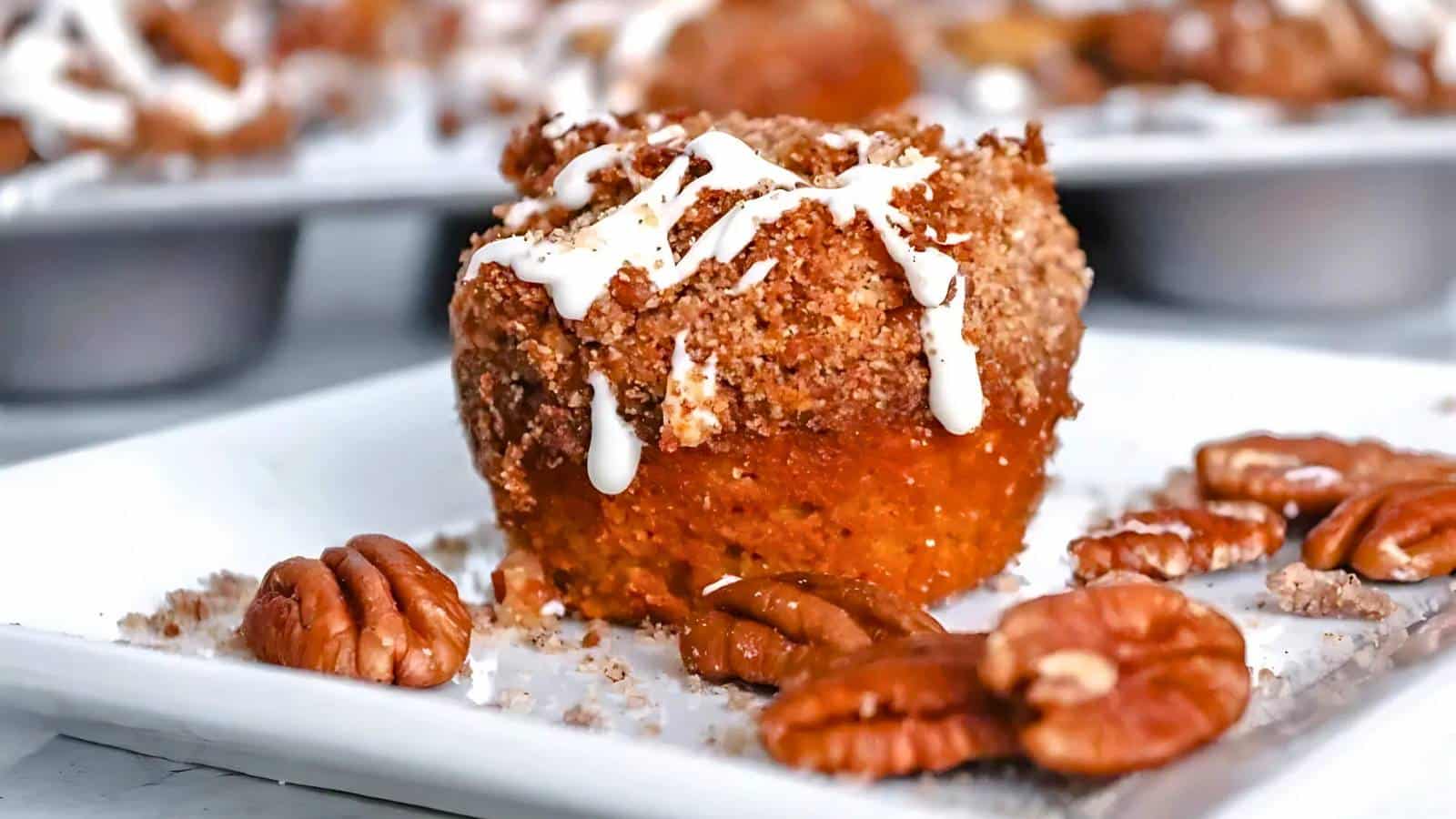 Close-up of a crumb muffin topped with icing on a white plate, surrounded by pecans. Other muffins are blurred in the background.