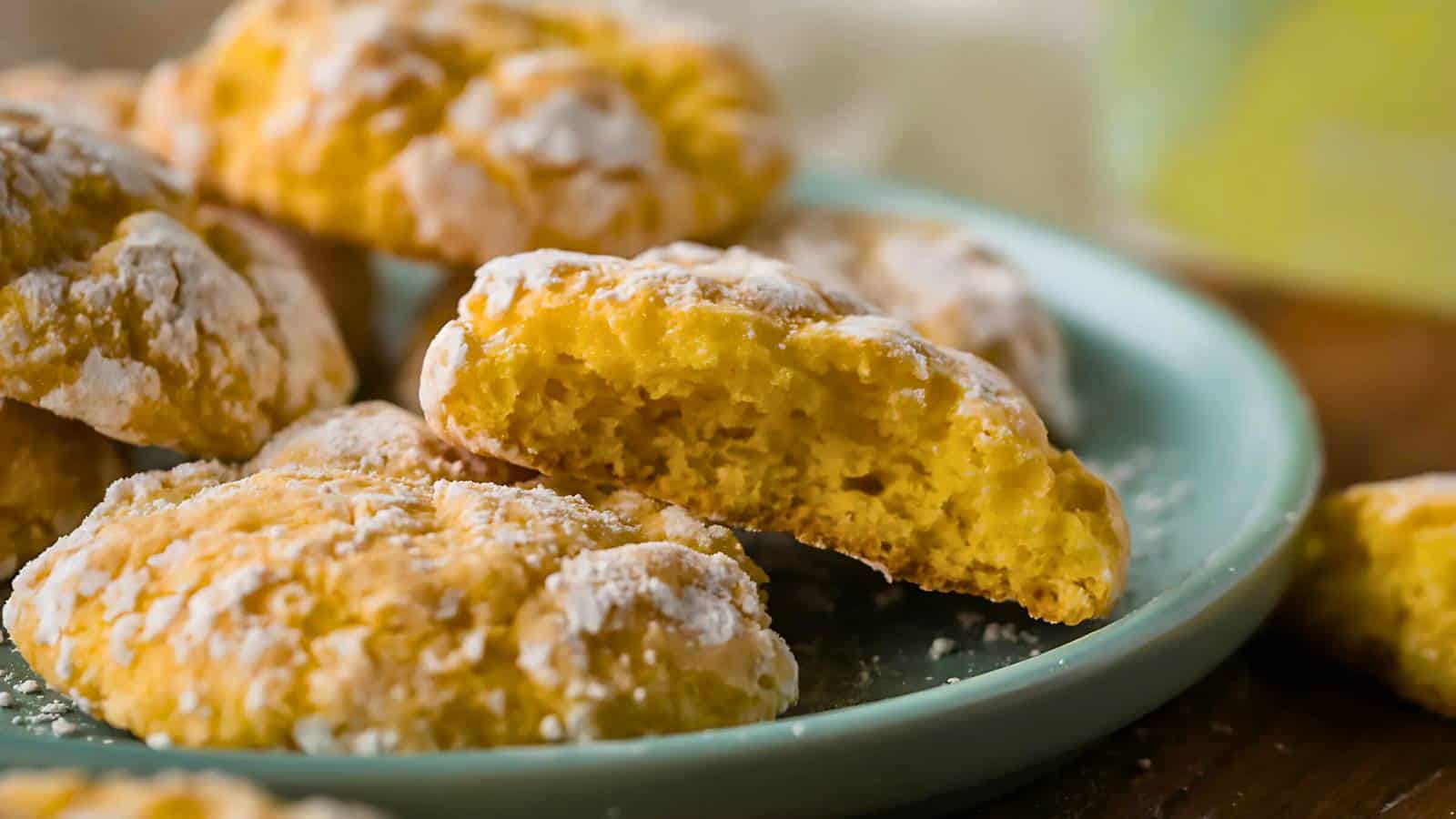 A plate of lemon crinkle cookies, with one broken in half to show the soft texture inside. Cookies are dusted with powdered sugar.