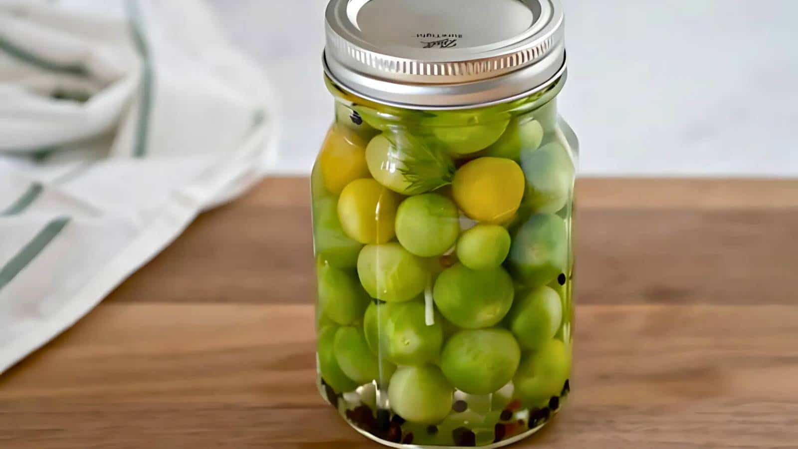 A jar of pickled green tomatoes with a metal lid sits on a wooden surface. A white cloth is partially visible in the background.