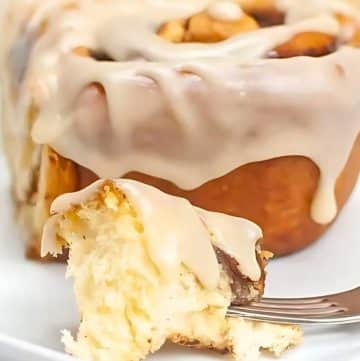 Close-up of a cinnamon roll with icing on a white plate, with a fork holding a bite-sized piece.