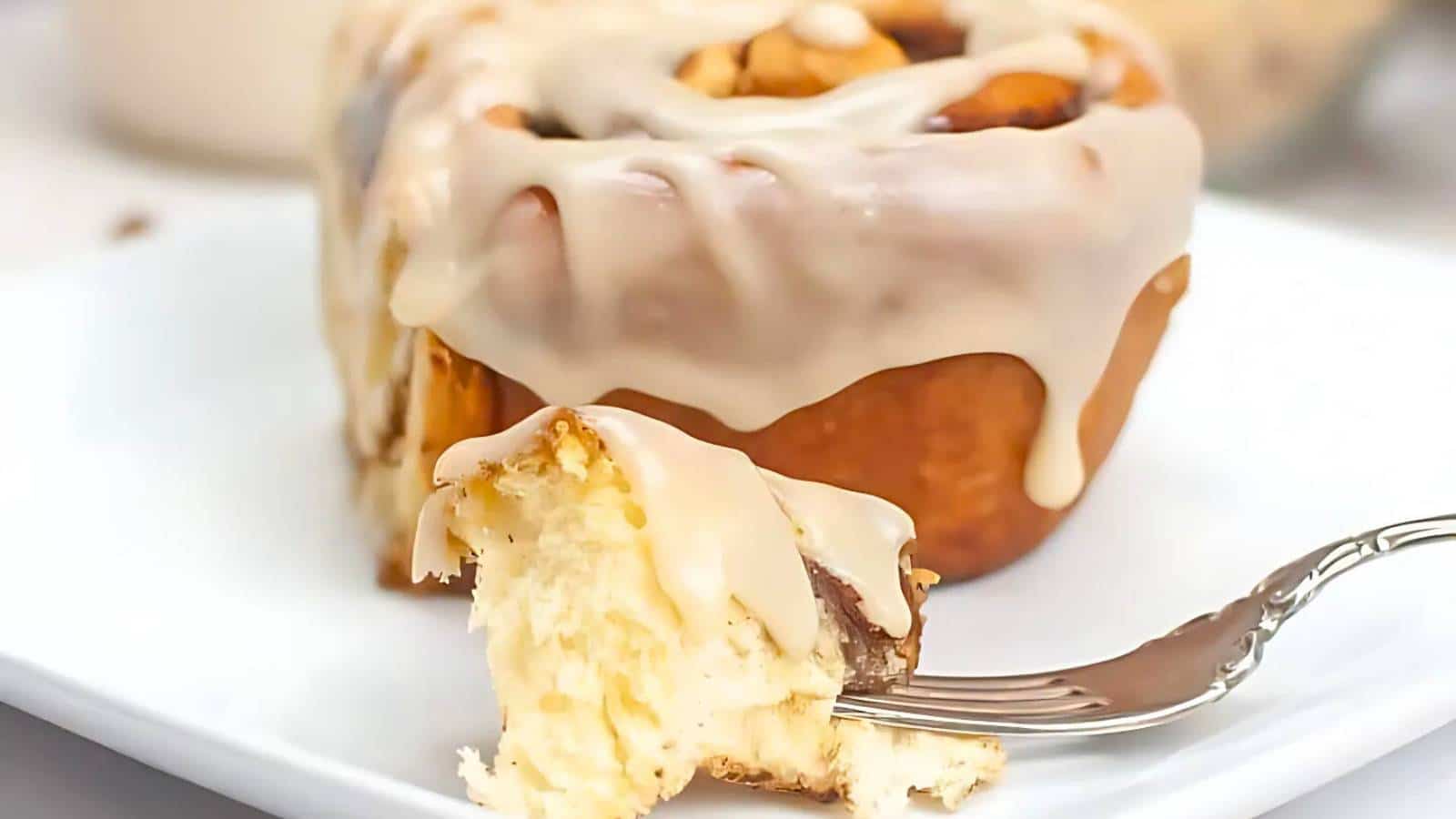 Close-up of a cinnamon roll with icing on a white plate, with a fork holding a bite-sized piece.