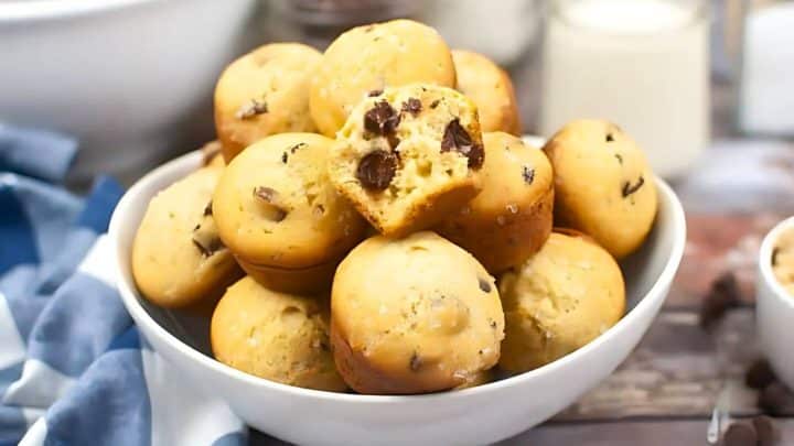 A bowl of chocolate chip muffins stacked on top of one another, with one muffin partially broken open—perfect as family picnic snacks. A napkin and a glass of milk are visible in the background.