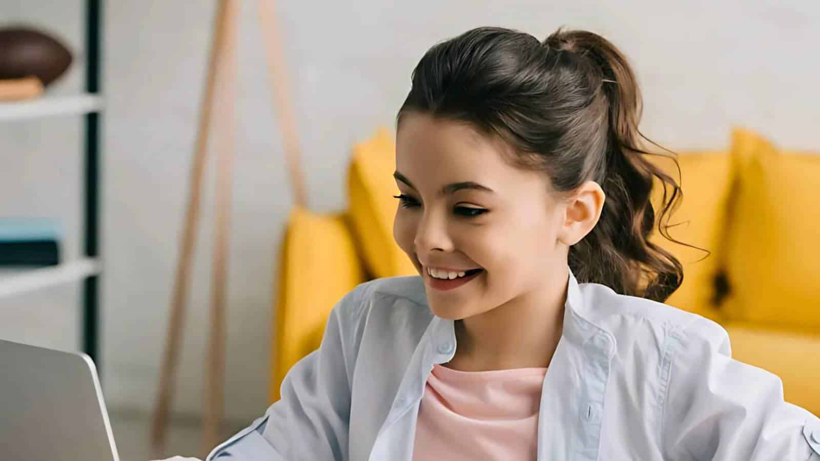A young girl with long dark hair in a ponytail smiles while looking at a laptop. She is wearing a white jacket over a pink shirt, sitting in a room with yellow decor.