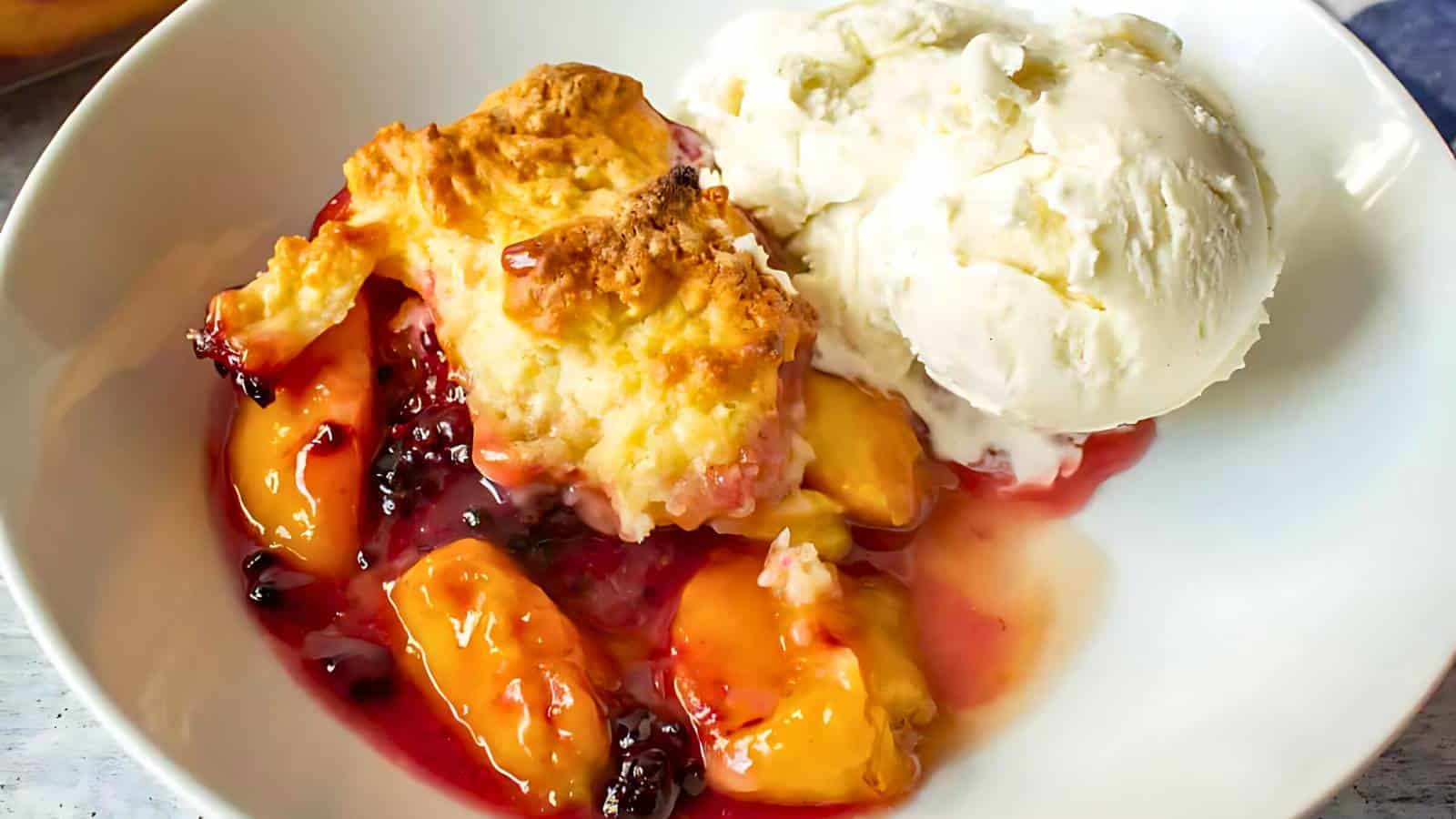 A white bowl containing peach and blackberry cobbler served with a scoop of vanilla ice cream.