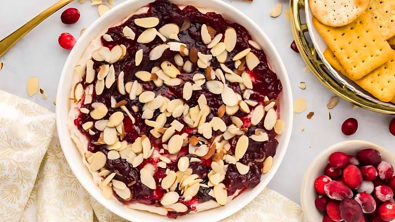 A baked brie topped with cranberry sauce and almond slices on a round dish, surrounded by crackers and fresh cranberries.
