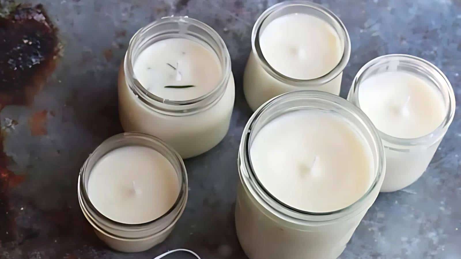 Five white candles in glass jars arranged on a textured gray surface.