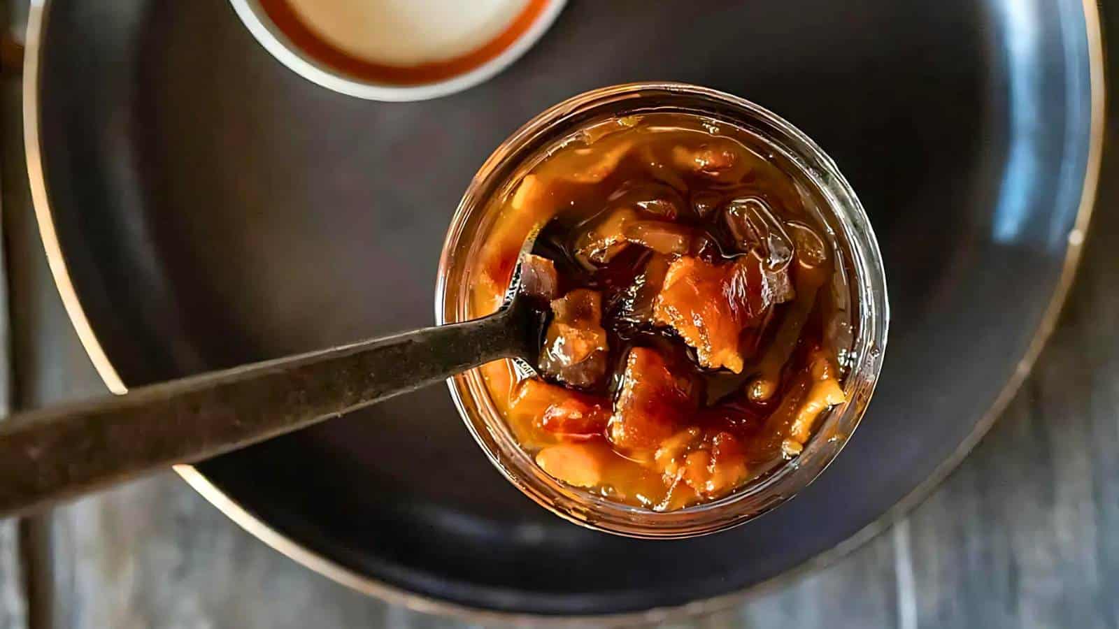 Overhead view of a jar filled with chutney, with a spoon inside, placed on a dark plate.