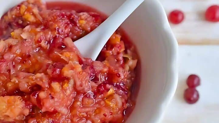 A white bowl filled with a red and orange chunky mixture, likely fruit-based, with a white spoon. Three small red fruits are scattered beside the bowl on a wooden surface.