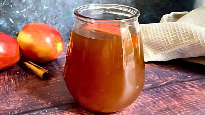 A glass of apple cider on a wooden table, with two apples, a cinnamon stick, and a beige cloth in the background.