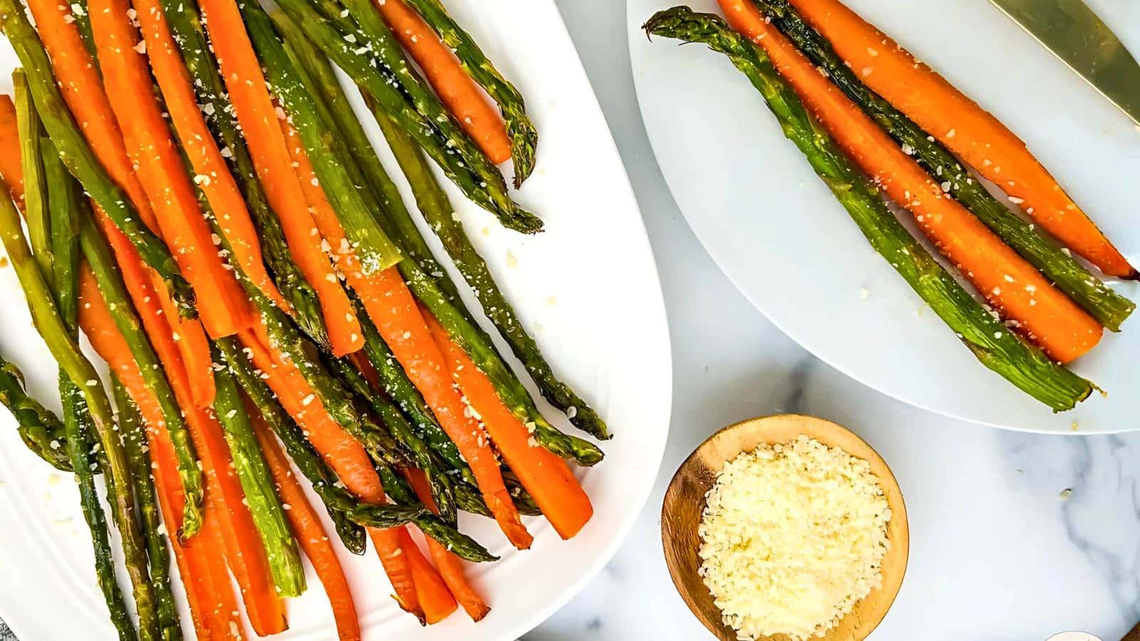 A white plate of roasted asparagus and carrots sprinkled with grated Parmesan cheese, a small wooden bowl containing more grated cheese, and another plate with the same vegetables and a knife.