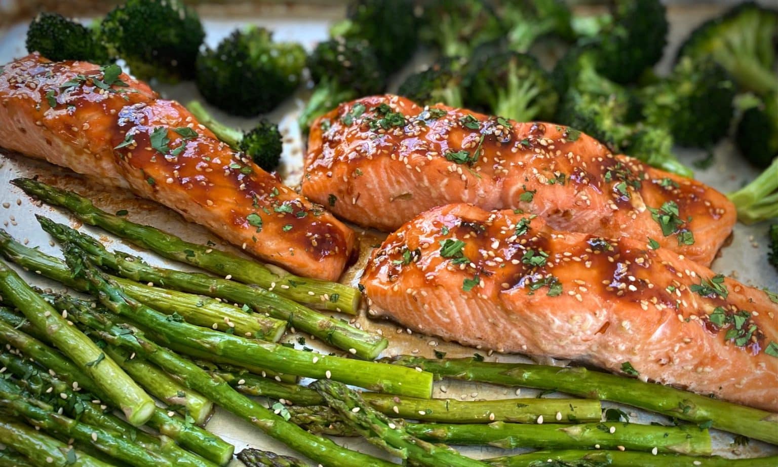 Baked salmon fillets glazed with sauce and garnished with herbs, served with roasted asparagus and broccoli on a tray.