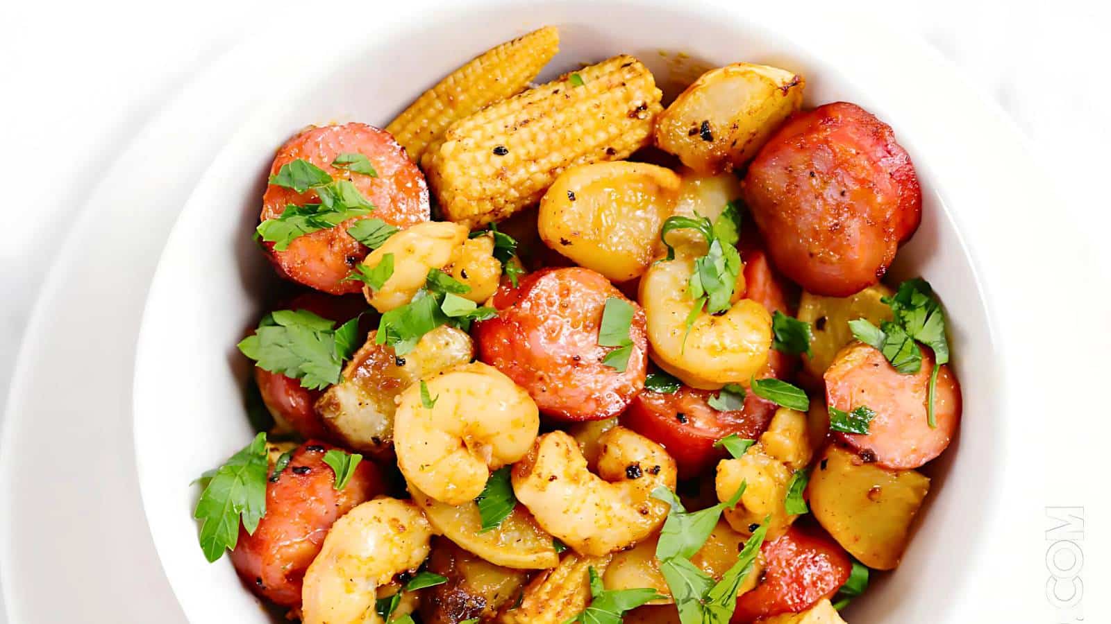 A bowl of shrimp, sausage, baby corn, and potatoes garnished with fresh parsley.