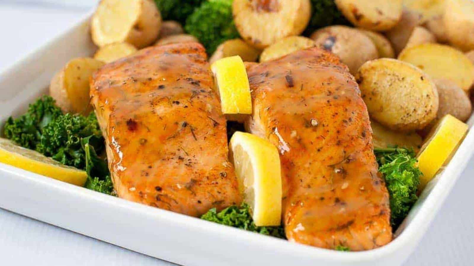 Grilled salmon fillets topped with sauce, garnished with lemon wedges, served on a bed of kale with roasted potatoes in a white dish.