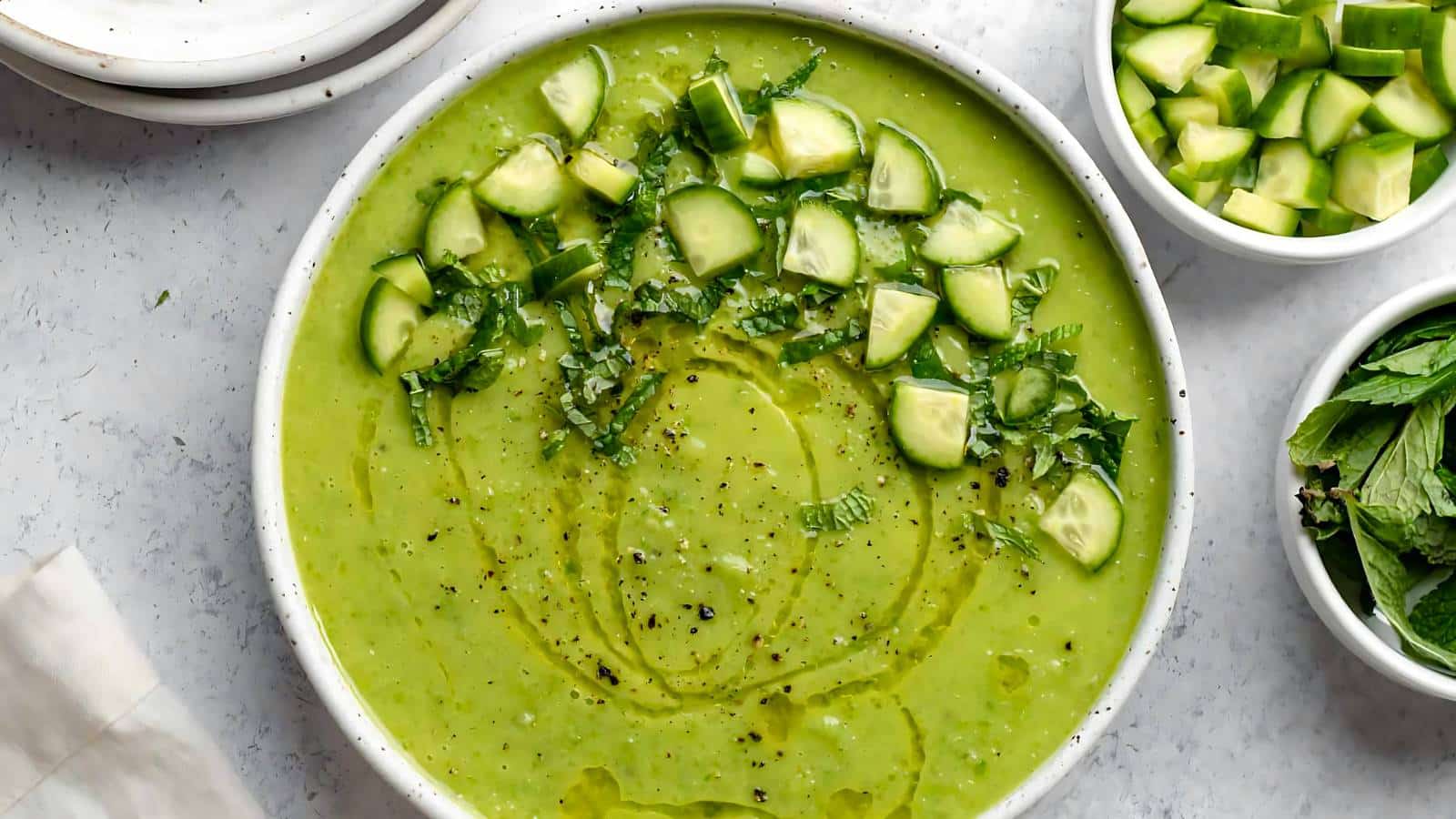 A bowl of creamy green soup garnished with cucumber chunks, black pepper, and herbs, placed on a light surface beside a bowl of chopped cucumbers and a bowl of fresh herbs.