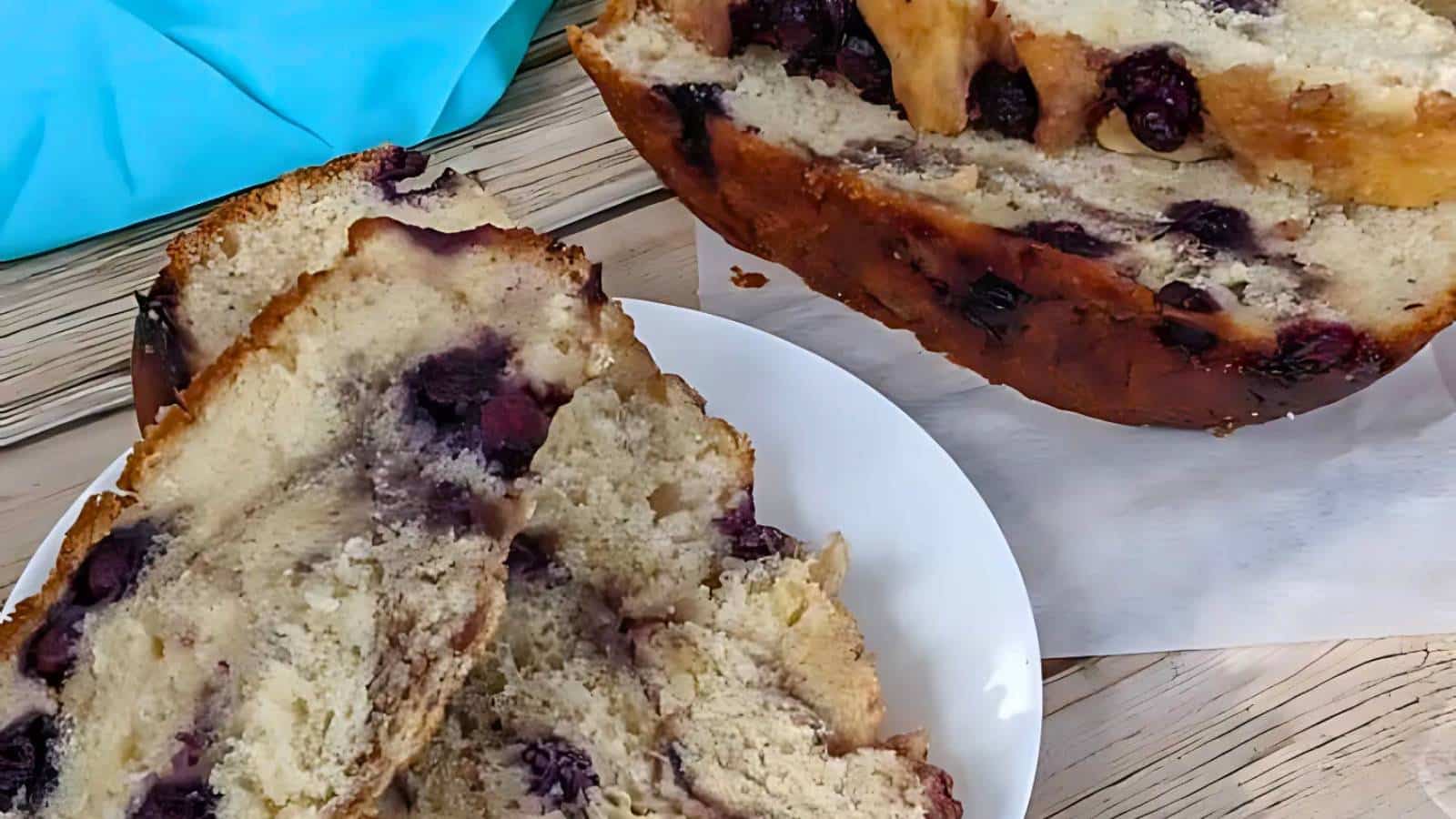 Sliced bread loaf with blueberries on a white plate, wooden table, and blue napkin in the background.