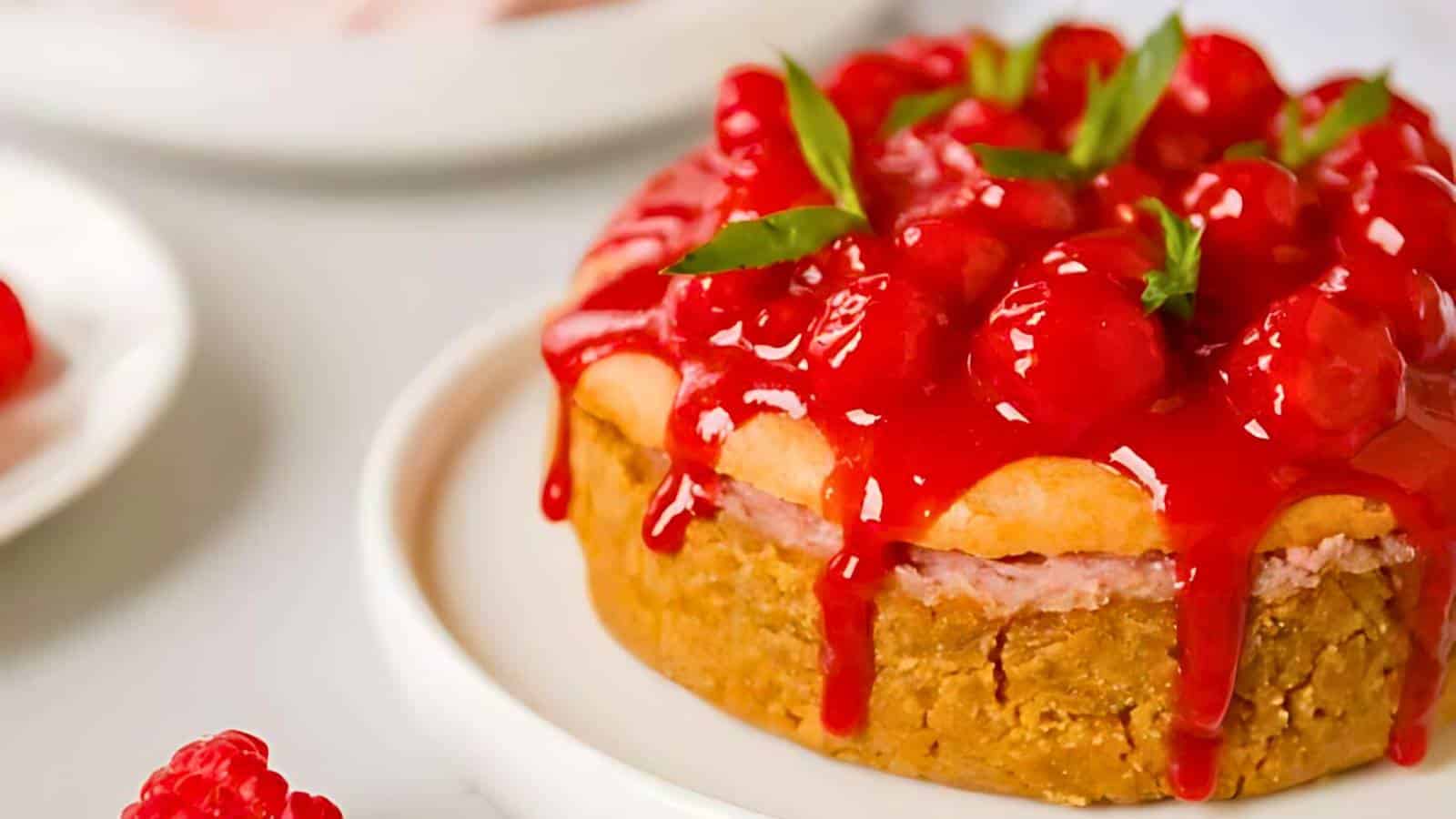 A small cheesecake topped with glossy red raspberries and garnished with green mint leaves, set on a white plate.