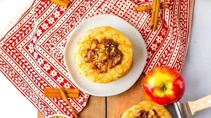 A pastry with apple filling is on a white plate, placed on a red and white patterned cloth. A whole apple, cinnamon sticks, and a knife are nearby.