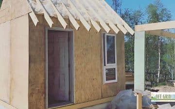 A small, unfinished wooden structure with an open door and window sits under construction outdoors. Surrounding trees and a banner advertising "Off Grid Life" are visible.