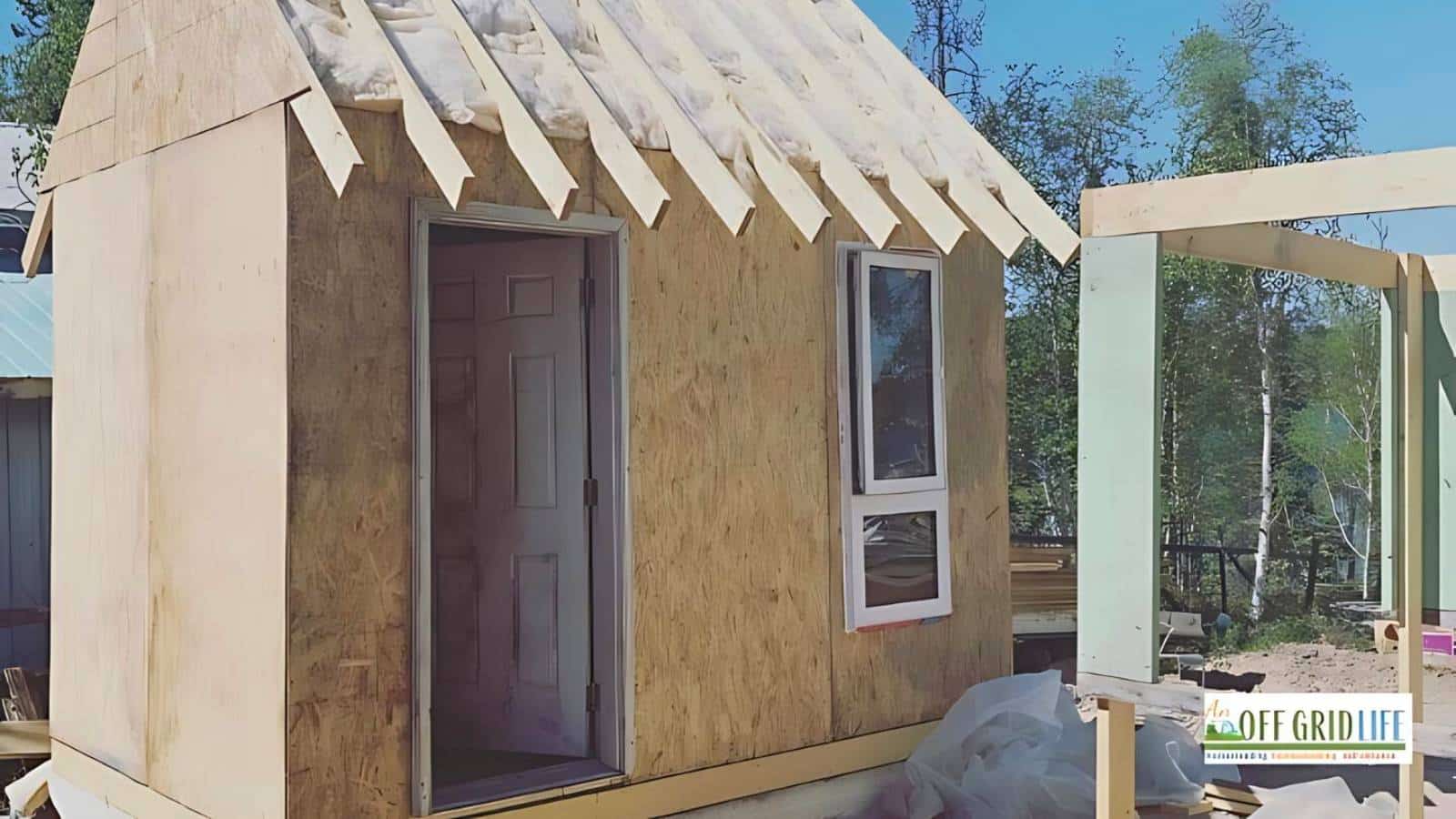 A small, unfinished wooden structure with an open door and window sits under construction outdoors. Surrounding trees and a banner advertising "Off Grid Life" are visible.