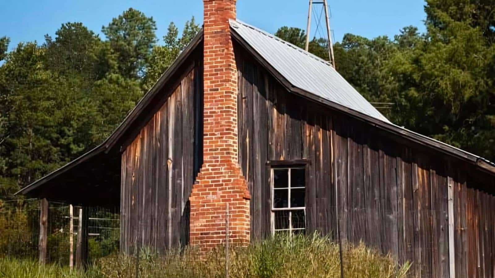 A rustic wooden cabin with a red brick chimney stands amidst tall grass and trees under a clear blue sky.
