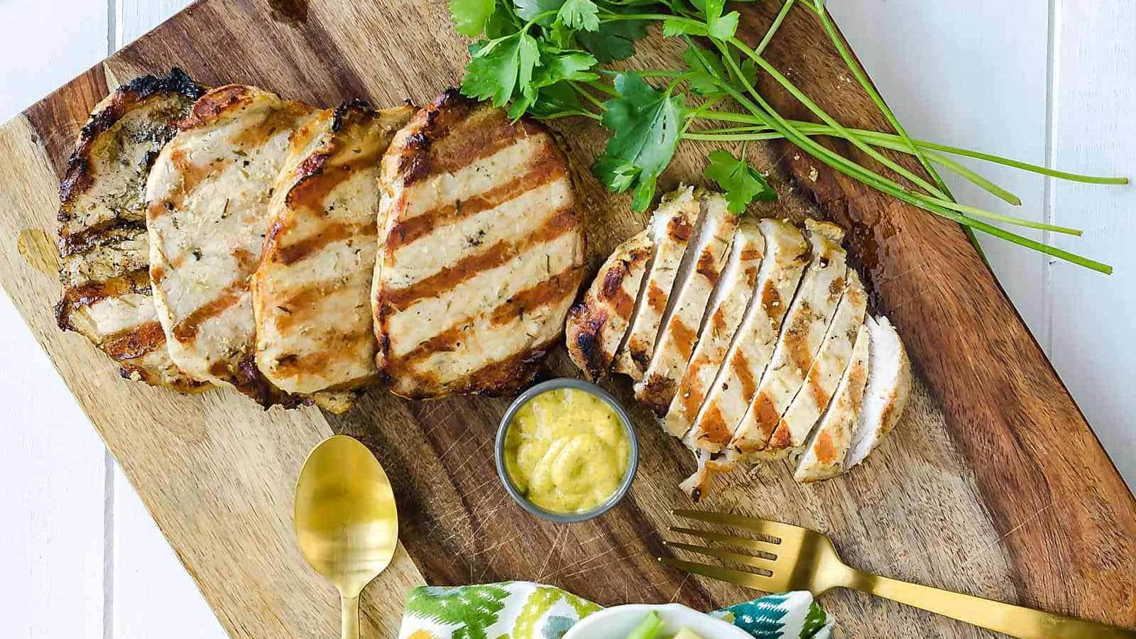 Grilled chicken breast slices on a wooden board with a small bowl of mustard, gold utensils, and parsley garnish.