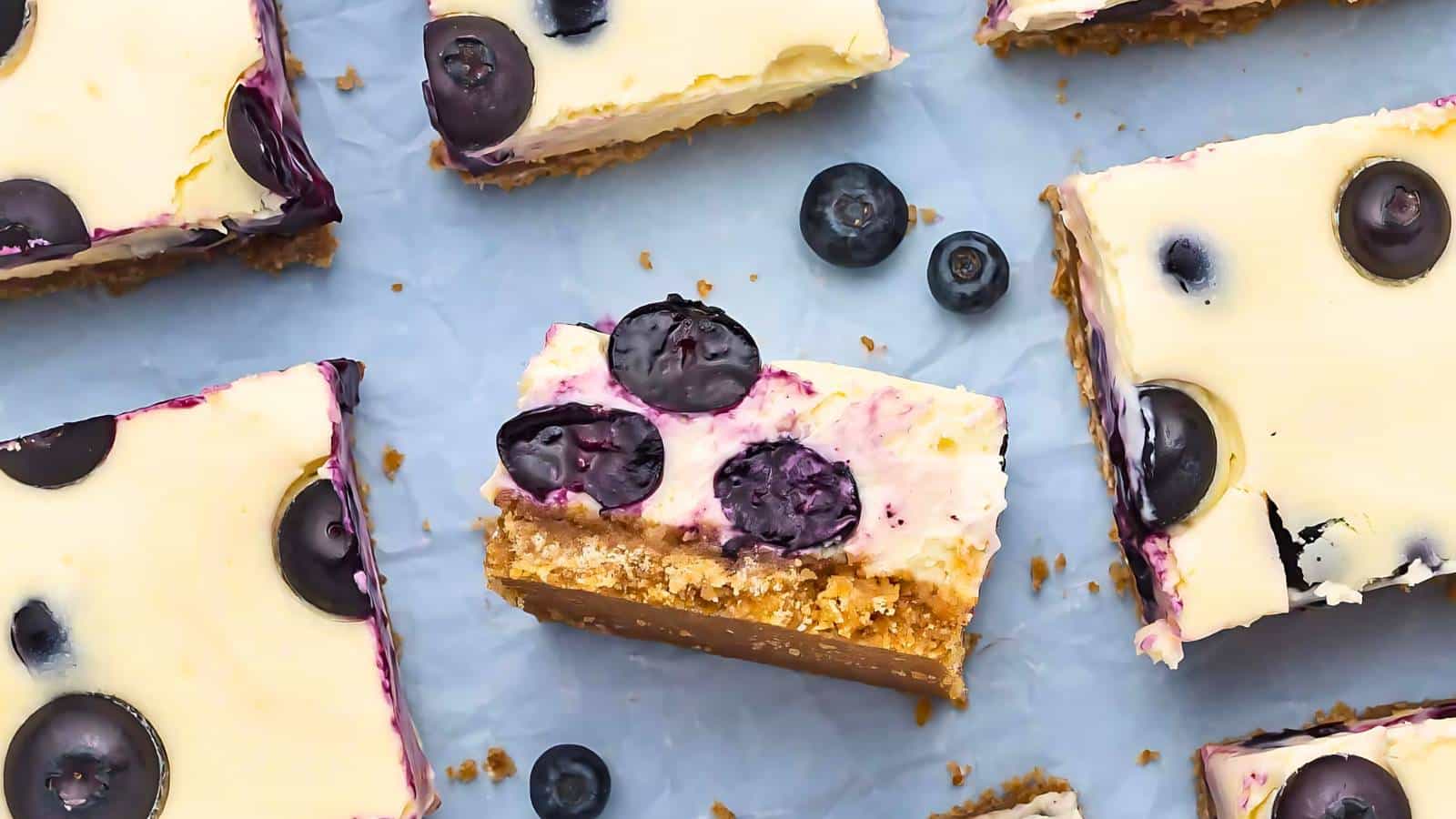 Blueberry cheesecake bars with crumbly crust are arranged on a parchment paper. Some bars are partially eaten and a few whole blueberries are visible around the bars.