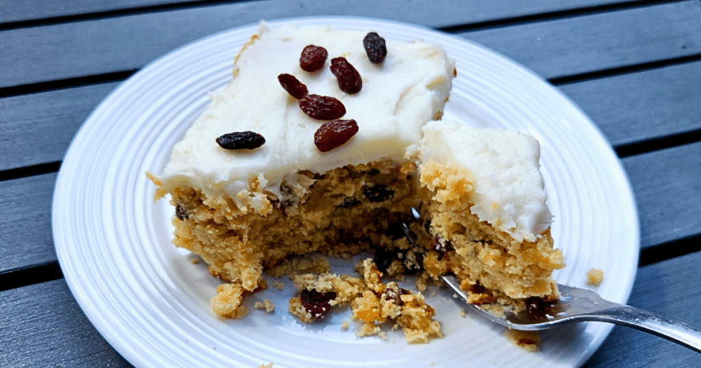 A slice of raisin cake with white frosting topped with raisins on a white plate. A fork has taken a piece out of the cake, revealing raisins inside. Plate is on a gray surface.