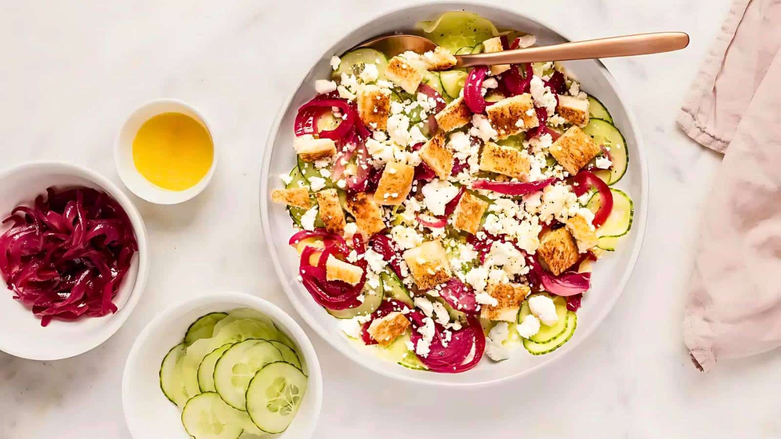 A fresh salad topped with croutons and feta cheese in a bowl, accompanied by bowls of dressing, sliced cucumbers, and pickled red onions on the side.