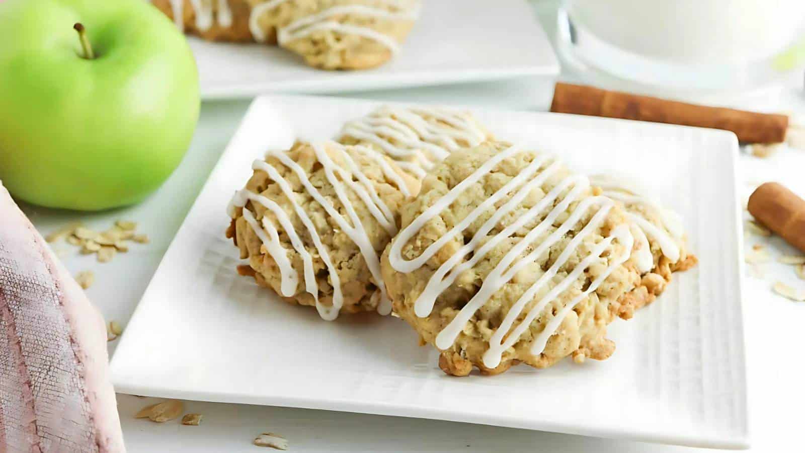 Two oatmeal cookies drizzled with white icing on a square white plate, with a green apple and cinnamon sticks in the background.