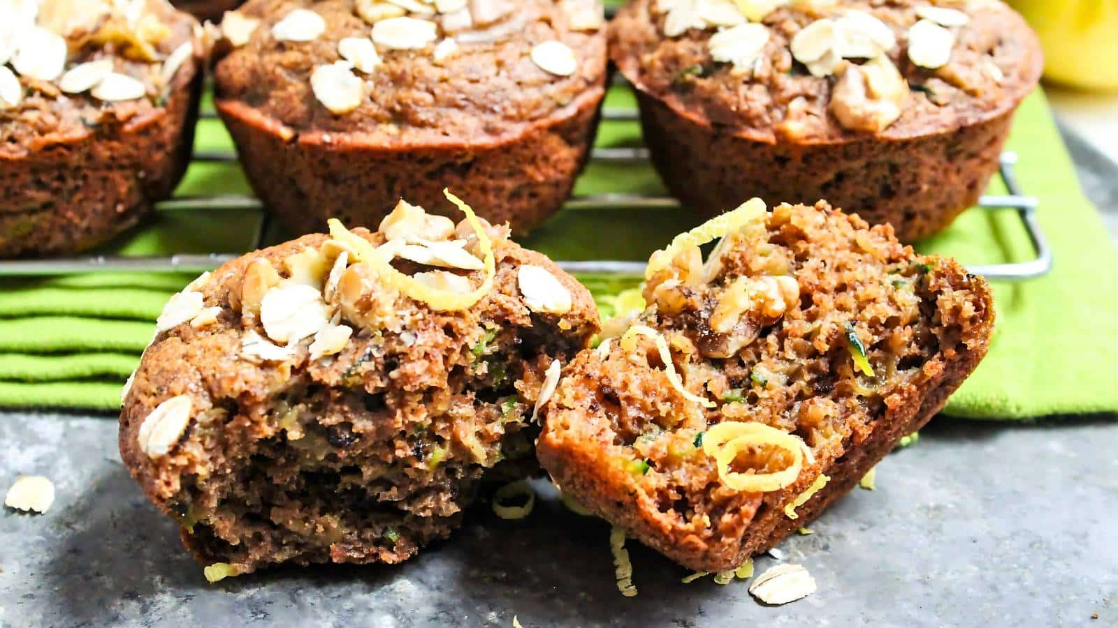 Freshly baked muffins with oats and nuts on top, one muffin halved, placed on a cooling rack with a green cloth underneath.