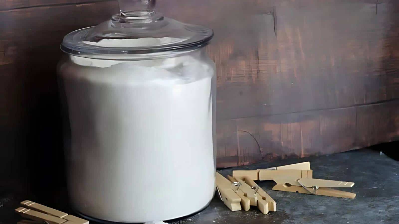 A large glass jar filled with white powder is placed on a dark surface. Several beige wooden clothespins are scattered nearby, against a wooden backdrop.