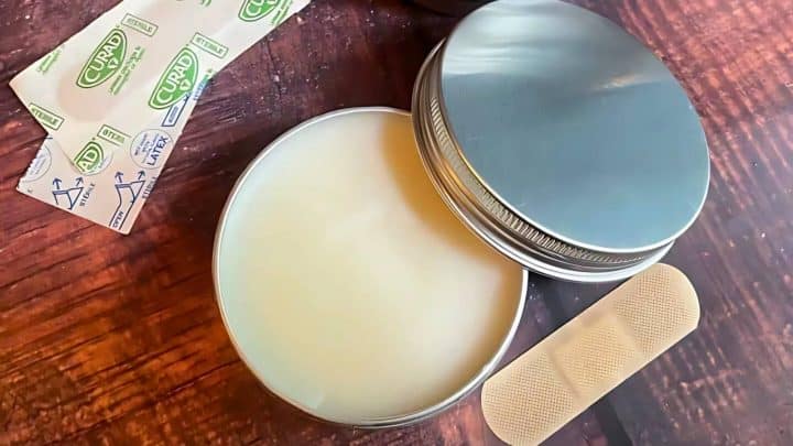 An open tin of balm on a wooden surface, surrounded by two band-aids and two packets labeled "CUARD.