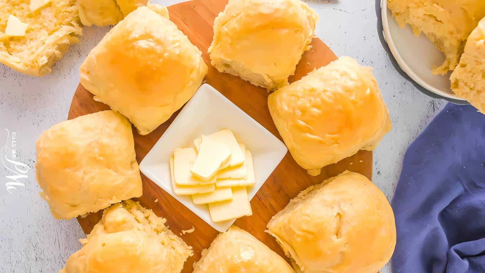 A wooden board with fluffy bread rolls encircling a square dish of sliced butter.