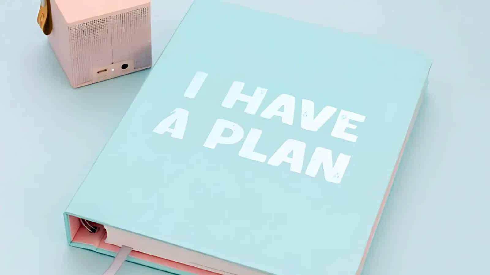 A light blue notebook with "I HAVE A PLAN" written on the cover, next to a small pink speaker, on a light blue surface.