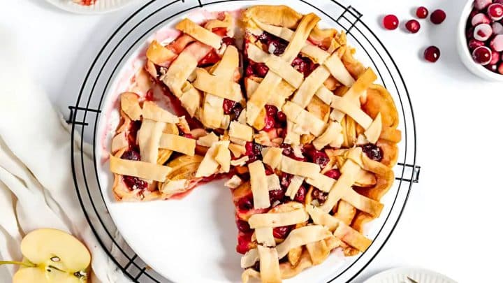 A lattice-topped pie with visible red and pink filling, possibly cherry or berry, is placed on a cooling rack. Sliced apples and whole cherries are nearby on a white surface.