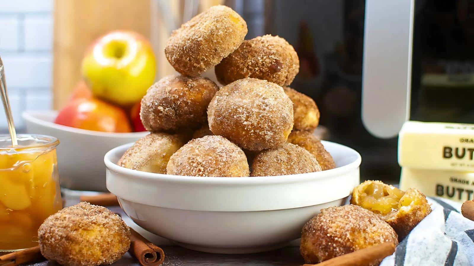 A white bowl filled with sugared doughnuts is surrounded by apples, butter sticks, cinnamon sticks, and a jar of apple sauce on a kitchen counter.