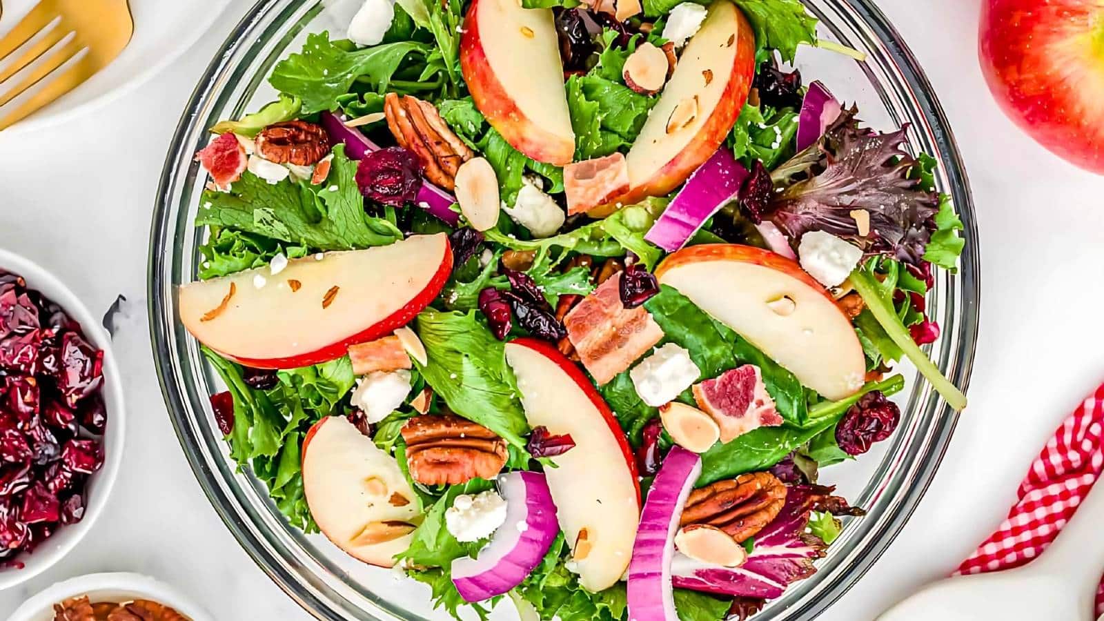 A salad with apple slices, pecans, red onion, goat cheese, bacon, and mixed greens in a glass bowl.